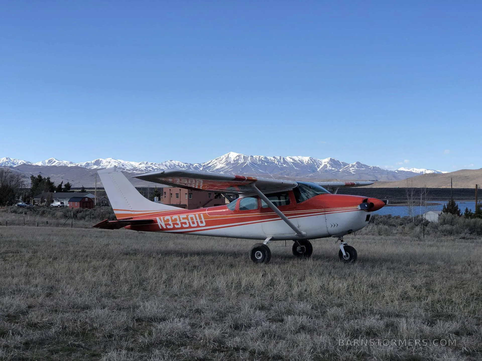 A red and white Cessna 182 Skylane (N3350U) is parked in a grassy field with snow-capped mountains and a lake in the background.