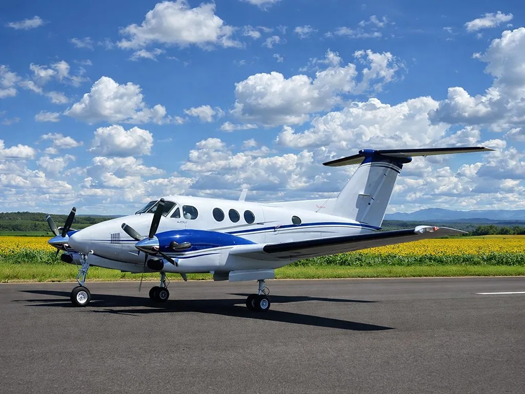 A white and blue twin-engine turboprop aircraft on a runway with a field of yellow flowers and blue sky in the background.
