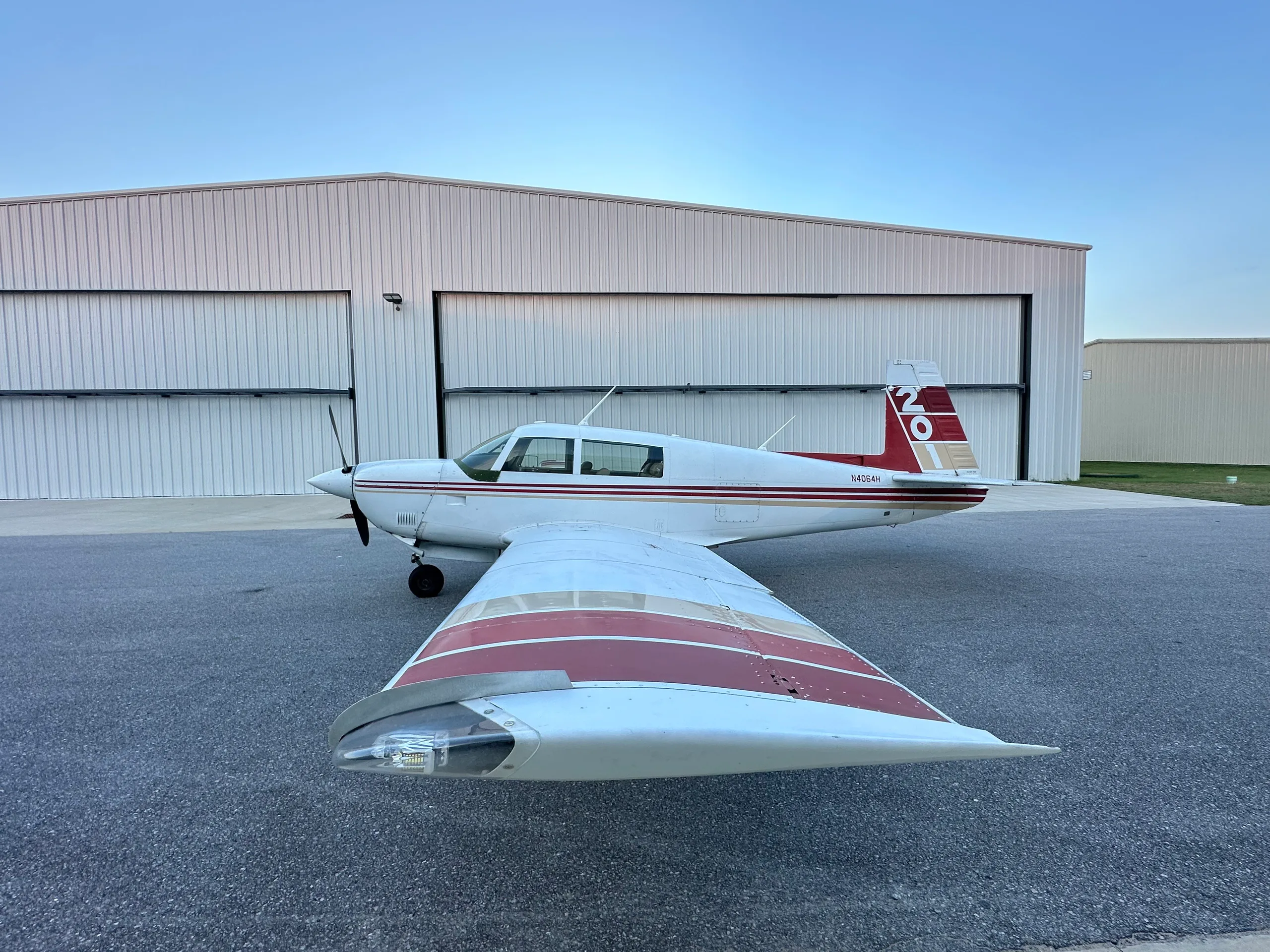 A white single-engine aircraft with red and gold stripes on the tail and wings, parked on an asphalt apron outside a hangar under a clear sky.