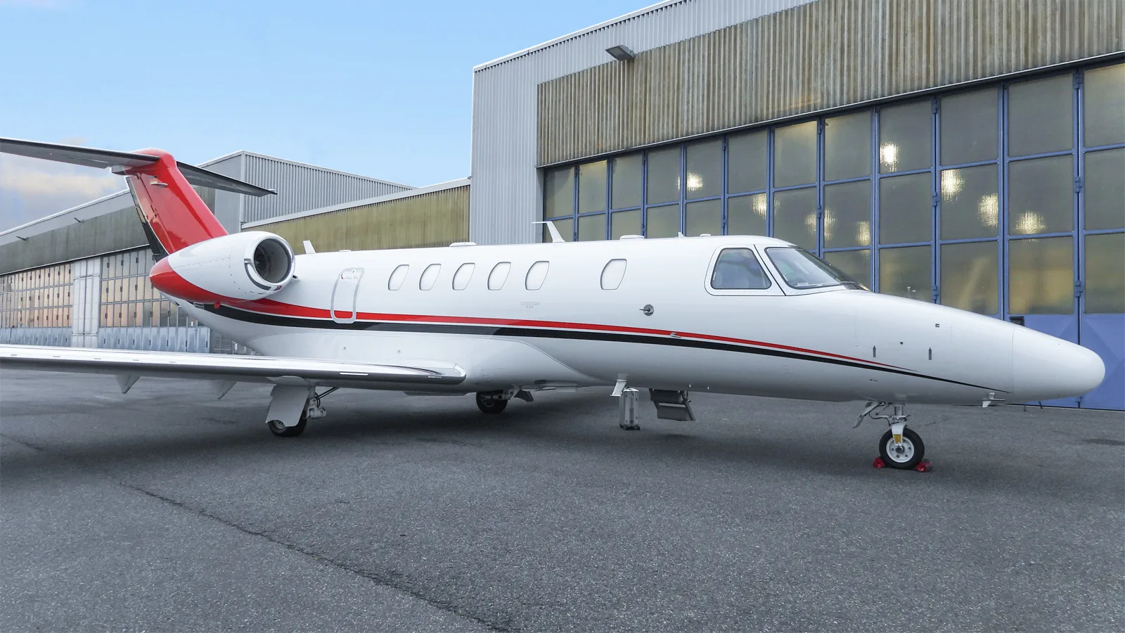 A full side view of a white Citation CJ4 business jet with red and black stripes parked on the tarmac in front of hangars.