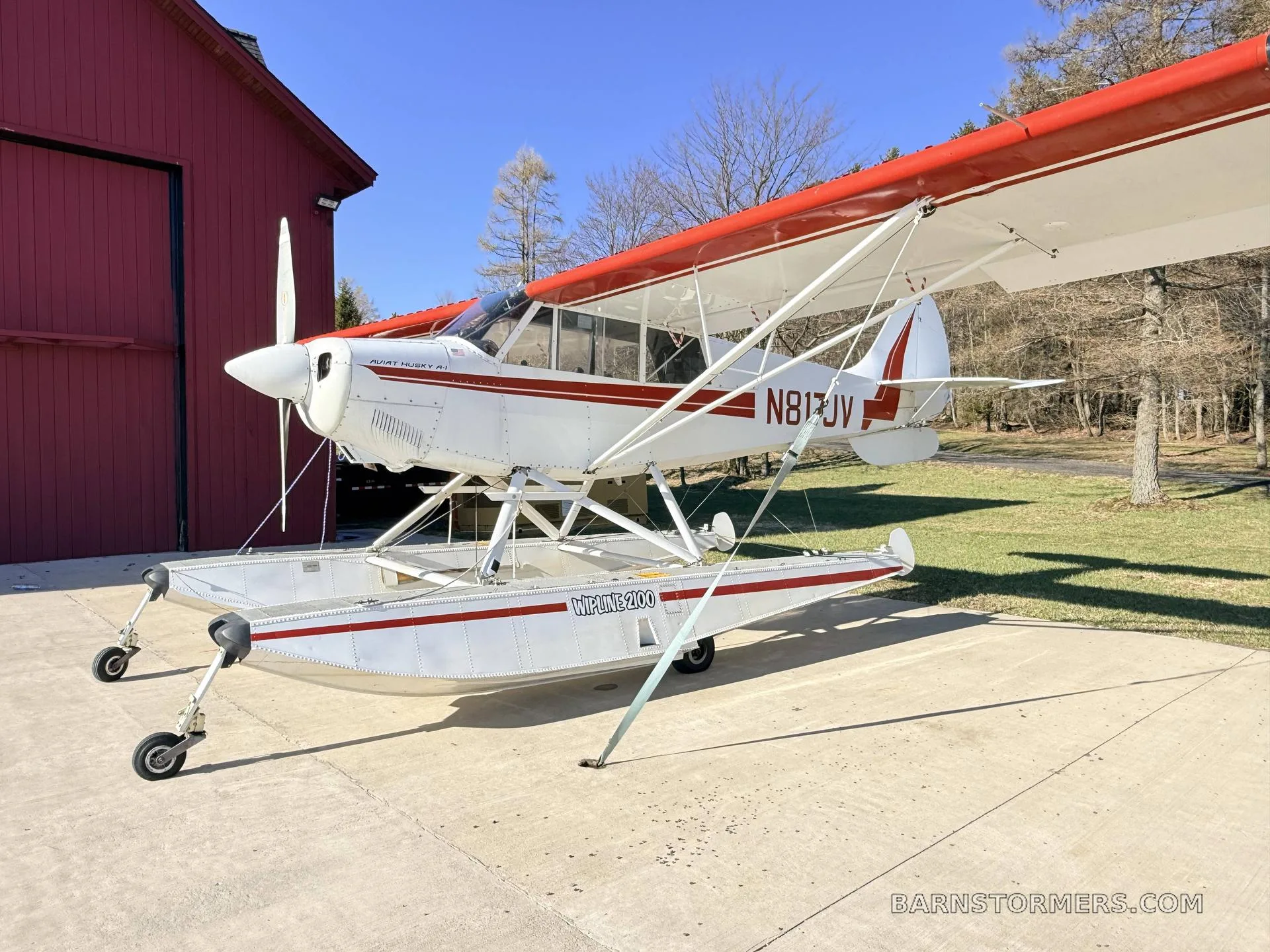 A white and red Aviat Husky A-1 floatplane (N817JV) parked next to a red hangar.