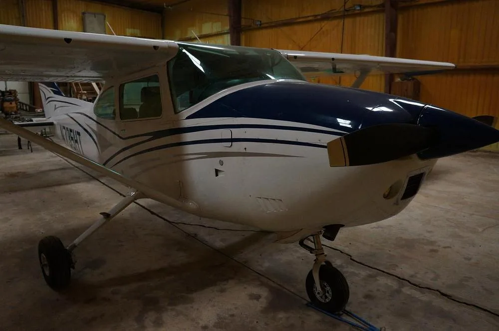 A white and blue Cessna 182 Skylane N735HT is parked in a hangar, viewed from the front.