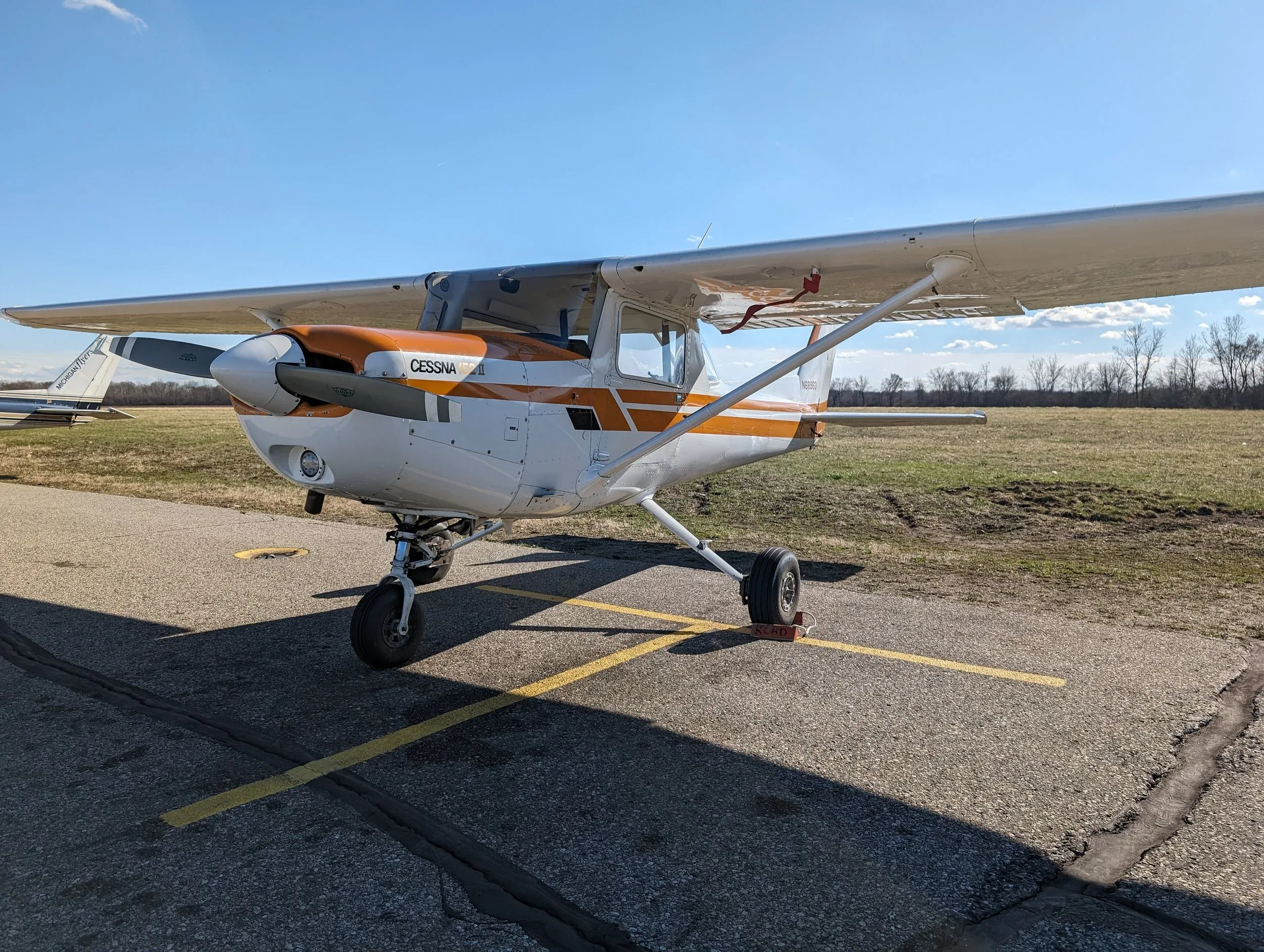 A white Cessna 172 aircraft with orange stripes is parked on an asphalt ramp under a clear blue sky.