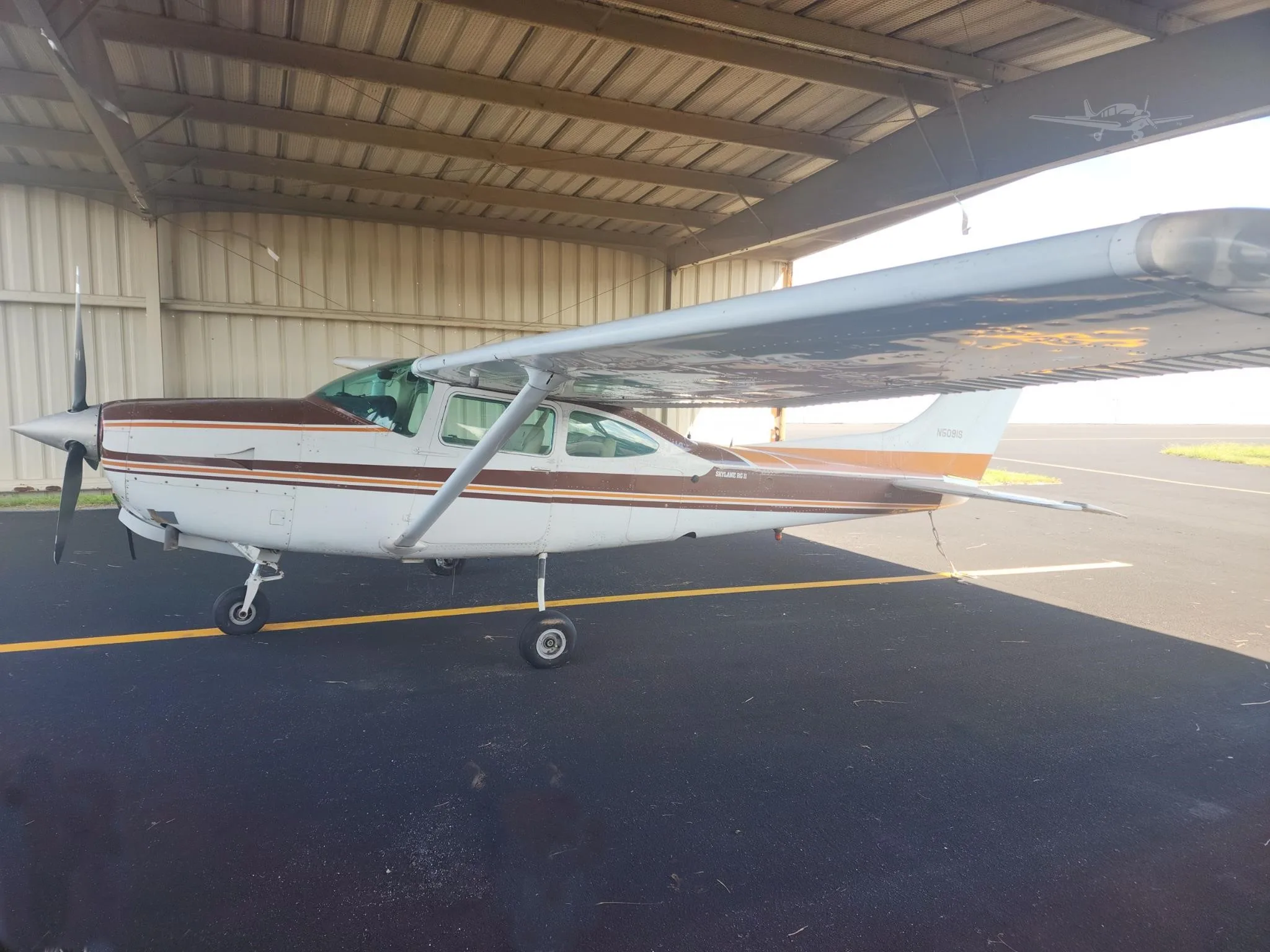 A white Cessna 182 Skylane II with brown and orange stripes is parked under a hangar roof on an asphalt ramp.