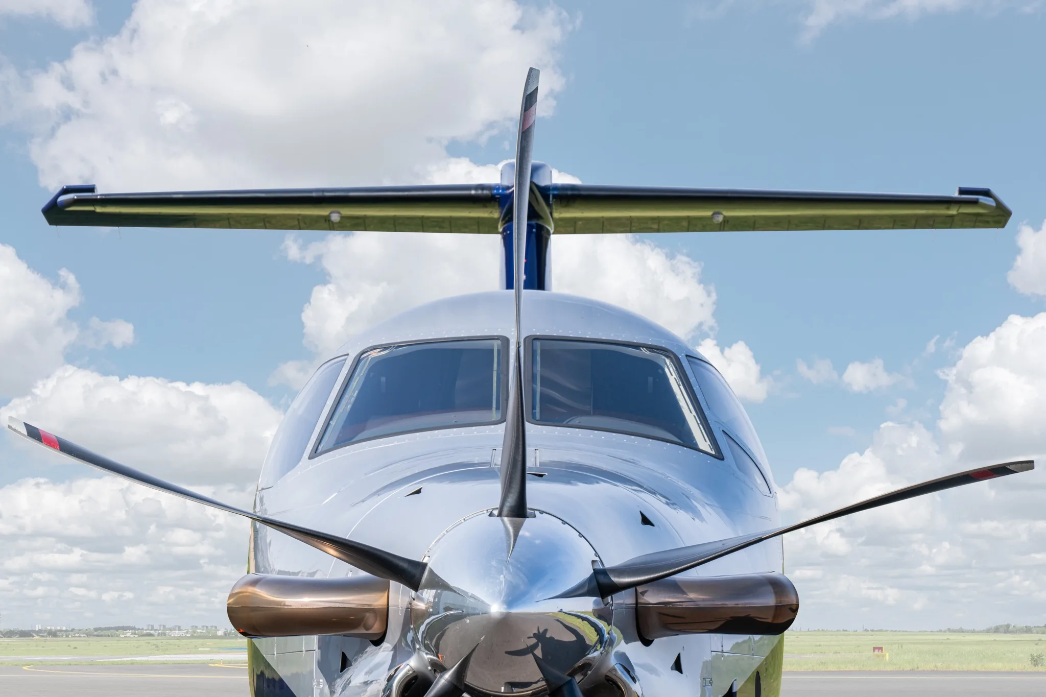 A head-on view of the silver Pilatus PC-12 NG aircraft with its propeller and cockpit against a cloudy sky.