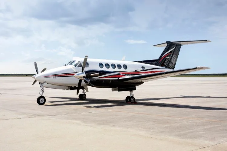 A white and black twin-turboprop aircraft with red stripes is parked on an airfield under a partly cloudy sky.