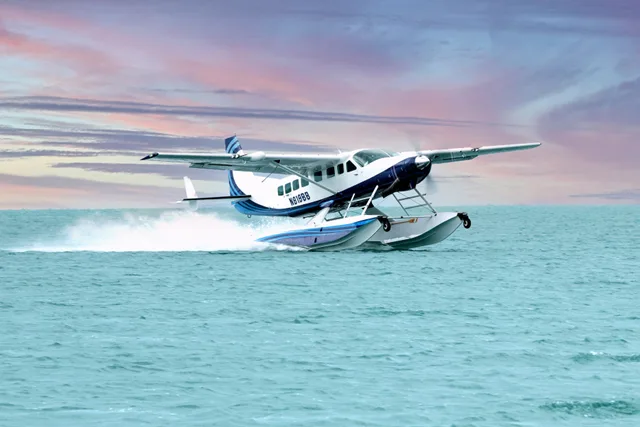 A white and blue Cessna Caravan seaplane with registration N819BB takes off from turquoise water, creating a spray, under a colorful sunset sky.