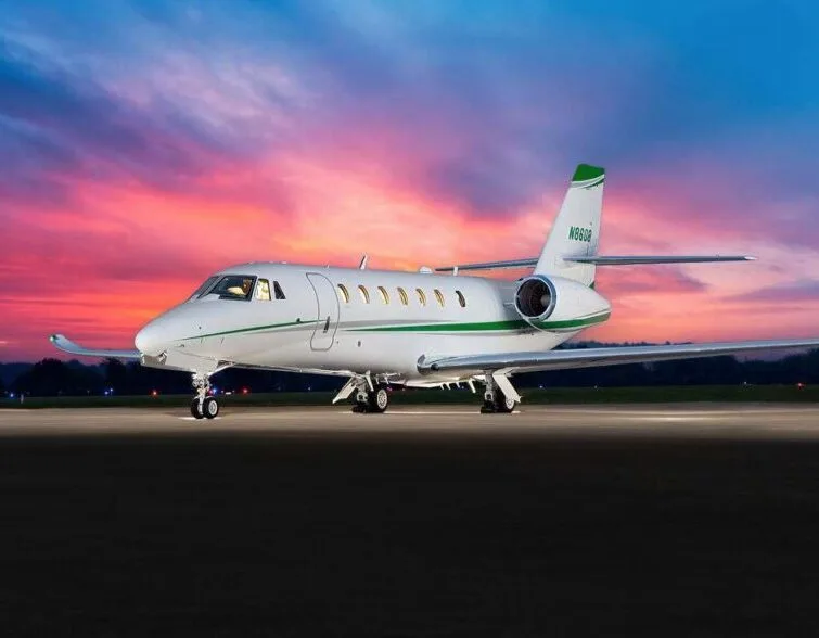 A white Cessna Citation X business jet with green accents is parked on a tarmac against a vibrant sunset sky.