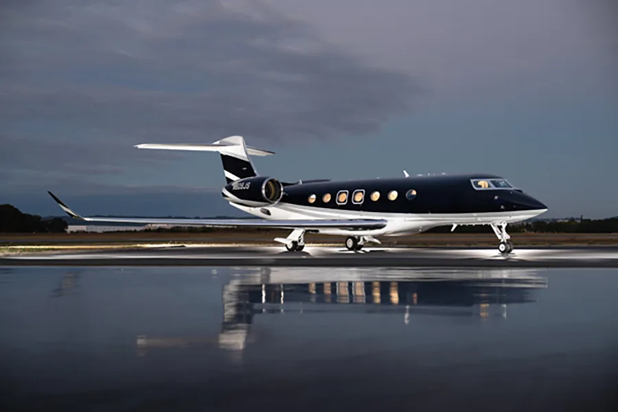 A dark blue and white Gulfstream G650 business jet, tail number N609JS, parked on a wet tarmac at dusk with reflections.