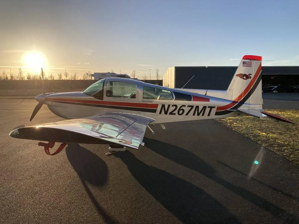 A silver, red, and black Grumman American AA-5B Tiger aircraft, N267MT, parked on an airfield at sunset.