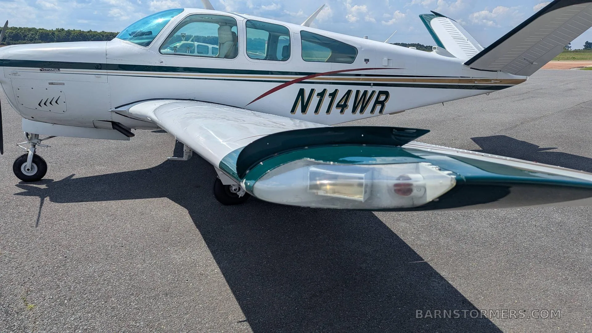 A full side view of the white Beechcraft Bonanza N114WR, with green and gold stripes, parked on an asphalt ramp.
