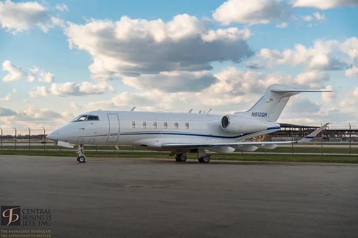 A white Bombardier Challenger business jet with a blue stripe is parked on an airfield under a partly cloudy sky.