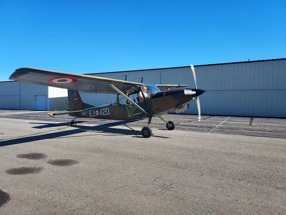 A side view of a single-engine, high-wing aircraft with olive drab paint and red wing markings, with its propeller spinning on an airfield.