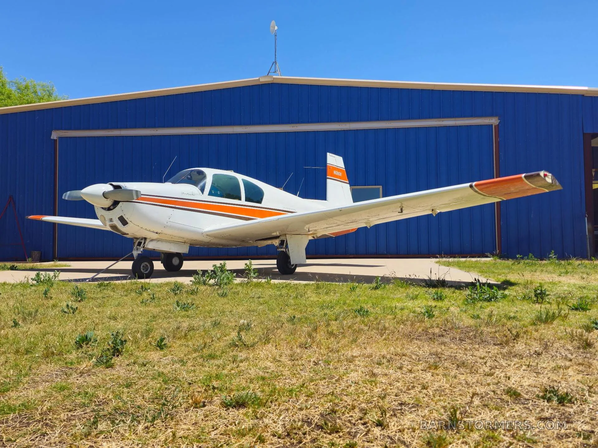 A white and orange single-engine aircraft (N282X) parked on grass in front of a blue hangar on a sunny day.