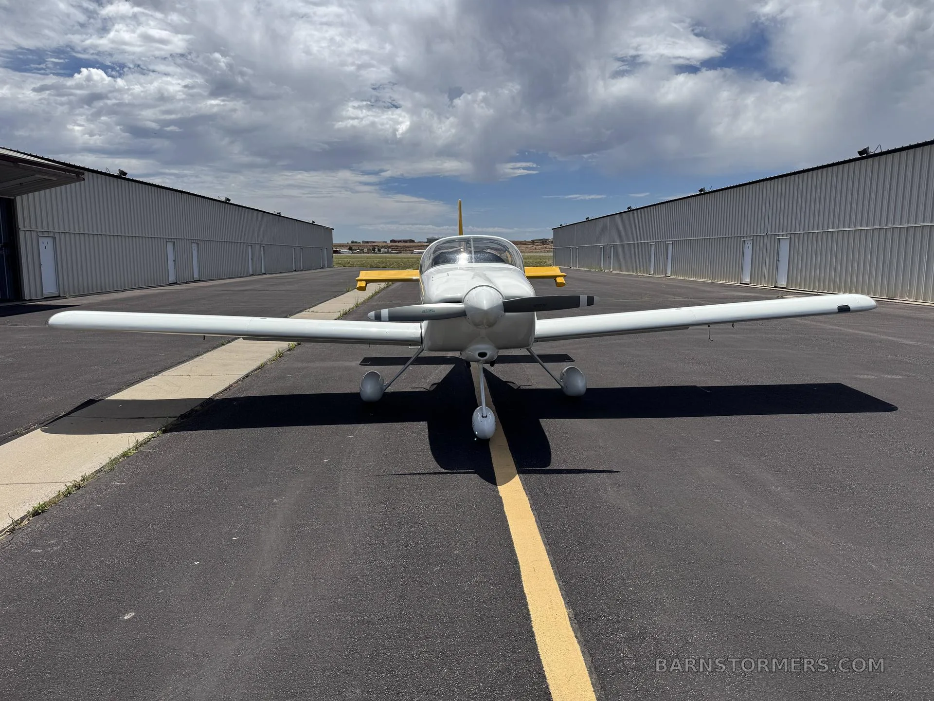 A front view of a white RV-9A aircraft with a yellow tail, parked on an asphalt runway with hangars on either side.