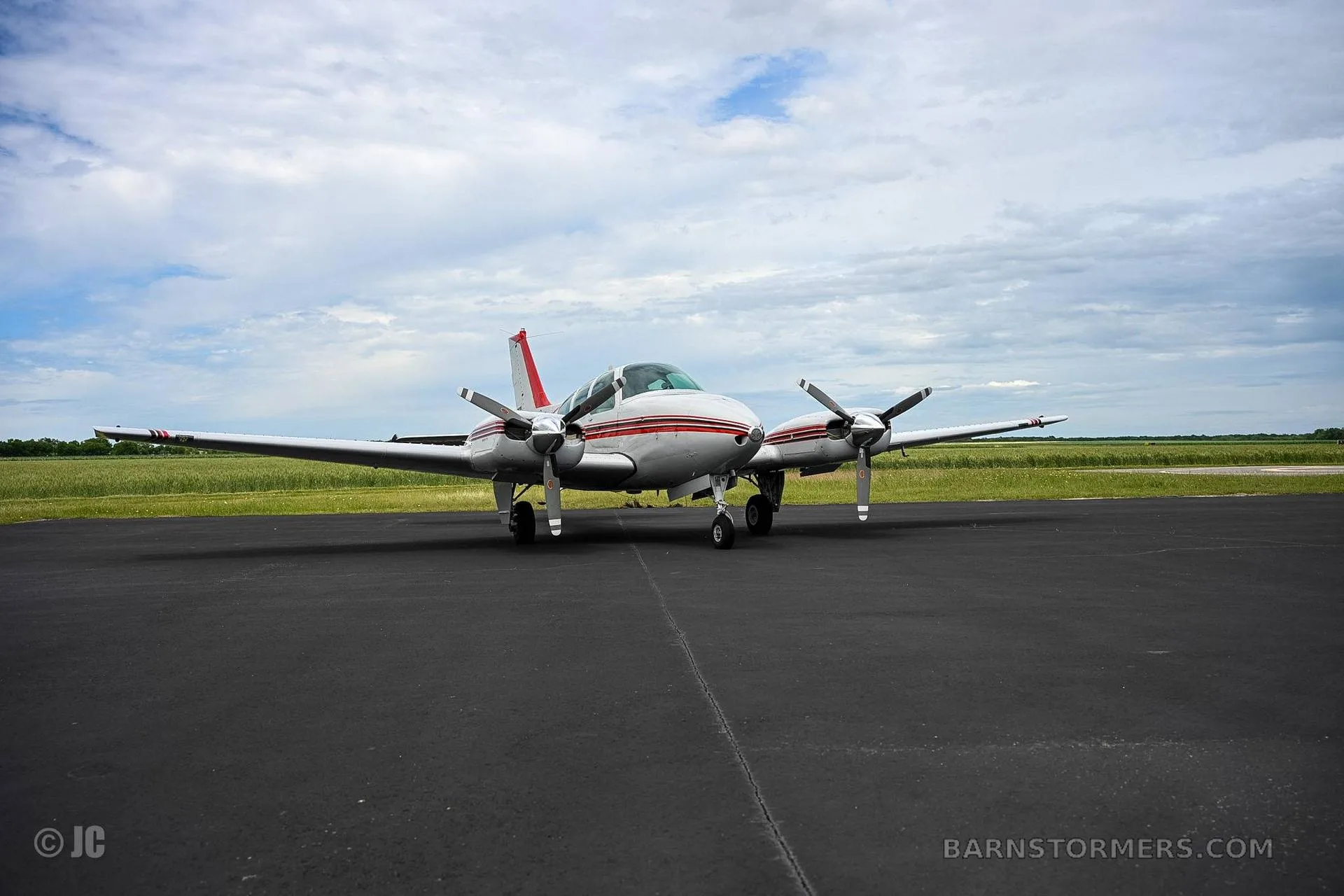 A wide shot of a white and red twin-engine propeller aircraft parked on an asphalt runway with green fields under a cloudy sky.