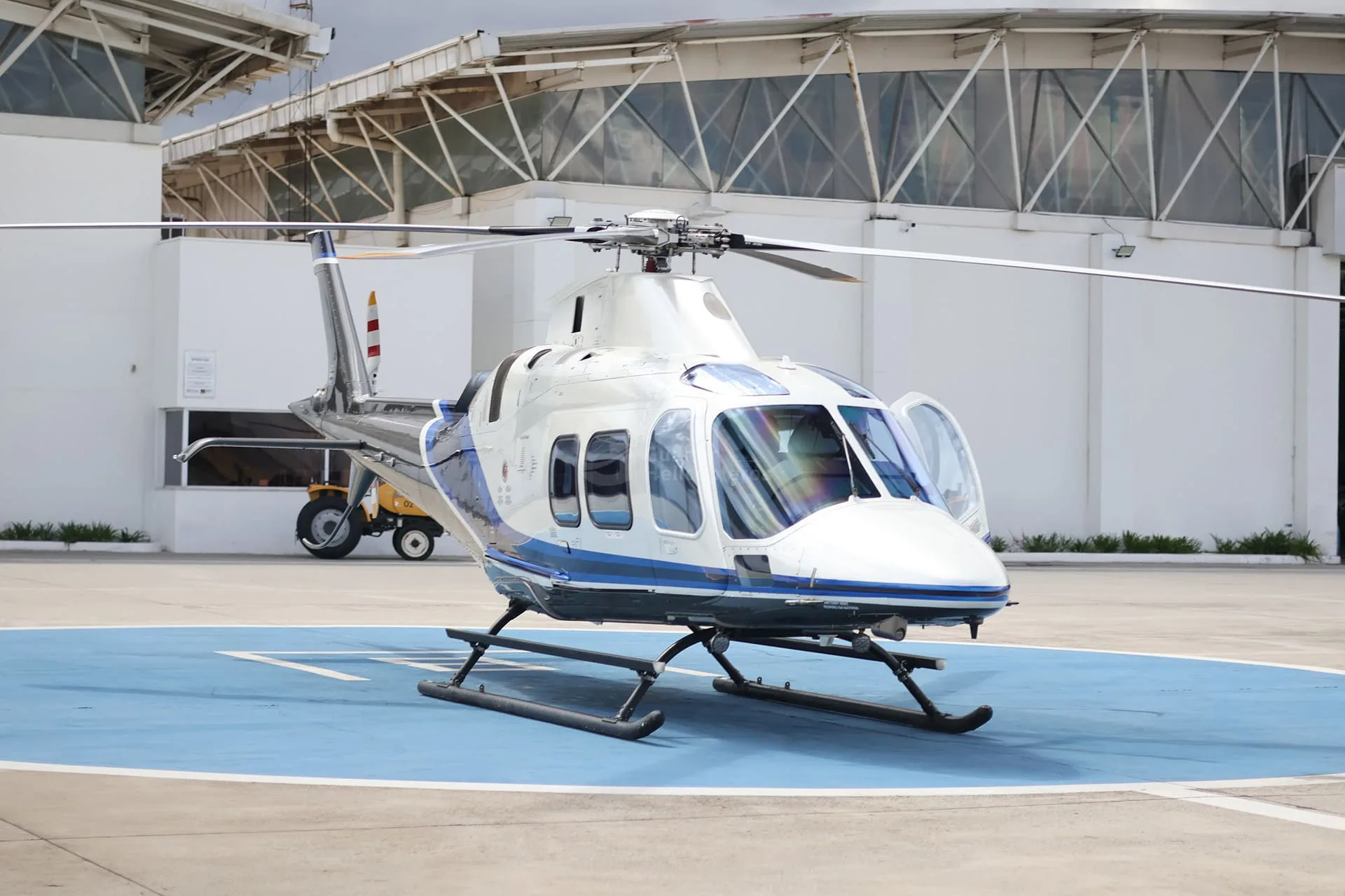A white and blue helicopter with both front doors open is parked on a blue helipad in front of a hangar.