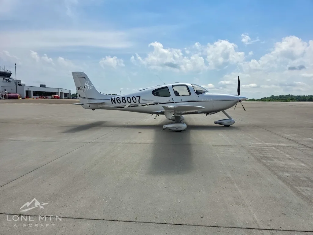 A silver Cirrus SR22 aircraft (N68007) with its doors closed, parked on an airfield with airport buildings in the background.