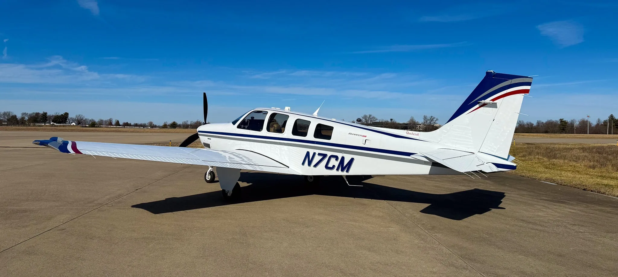 A white Beechcraft Bonanza G36 with blue and grey stripes, registration N7CM, parked on an airfield under a clear blue sky.