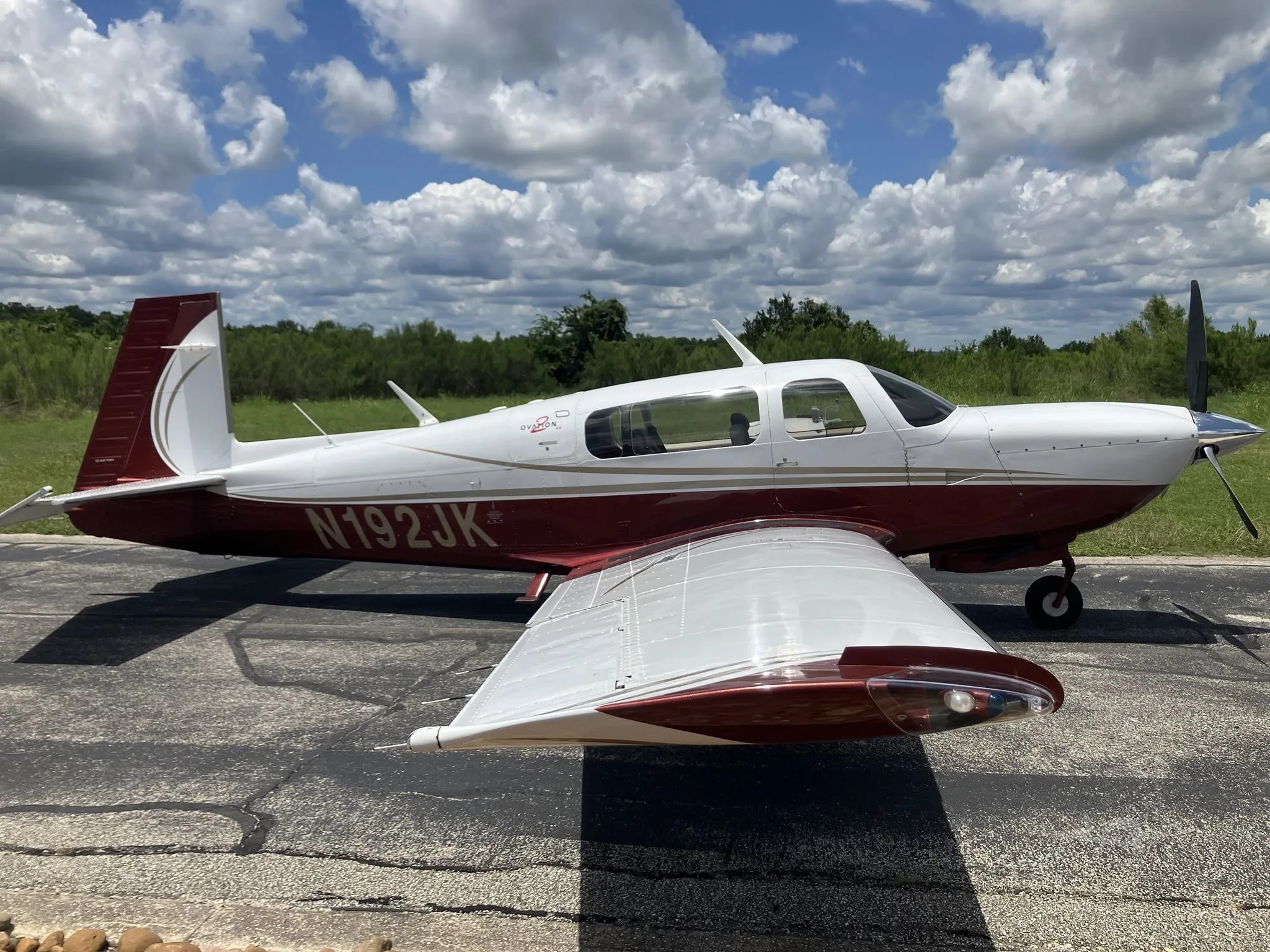 A white and maroon Mooney Ovation aircraft, registration N192JK, parked on an asphalt surface with a grassy background under a partly cloudy sky.