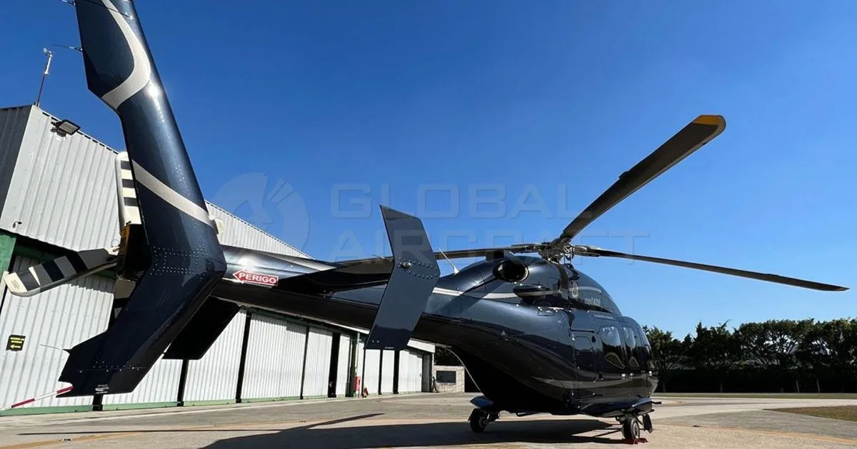 A dark blue Bell 429 helicopter with white stripes parked on an airfield next to a hangar, viewed from the rear-side.