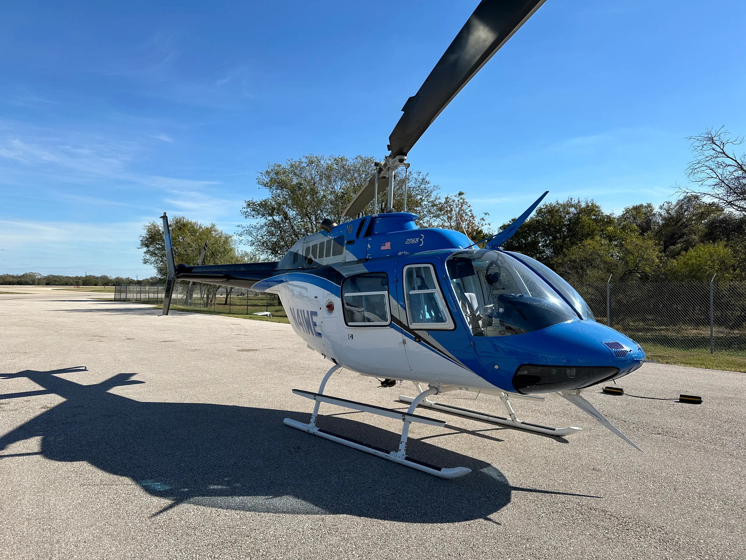 A blue and white Bell 206B3 helicopter with black accents is parked on an asphalt ramp under a clear blue sky.