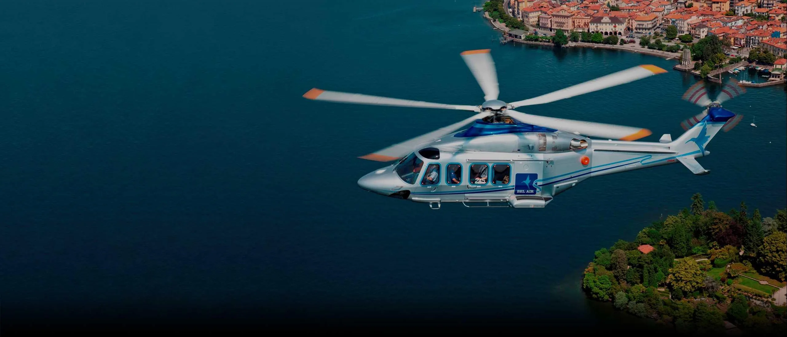 AgustaWestland AW169 helicopter flying over a lake near a town.