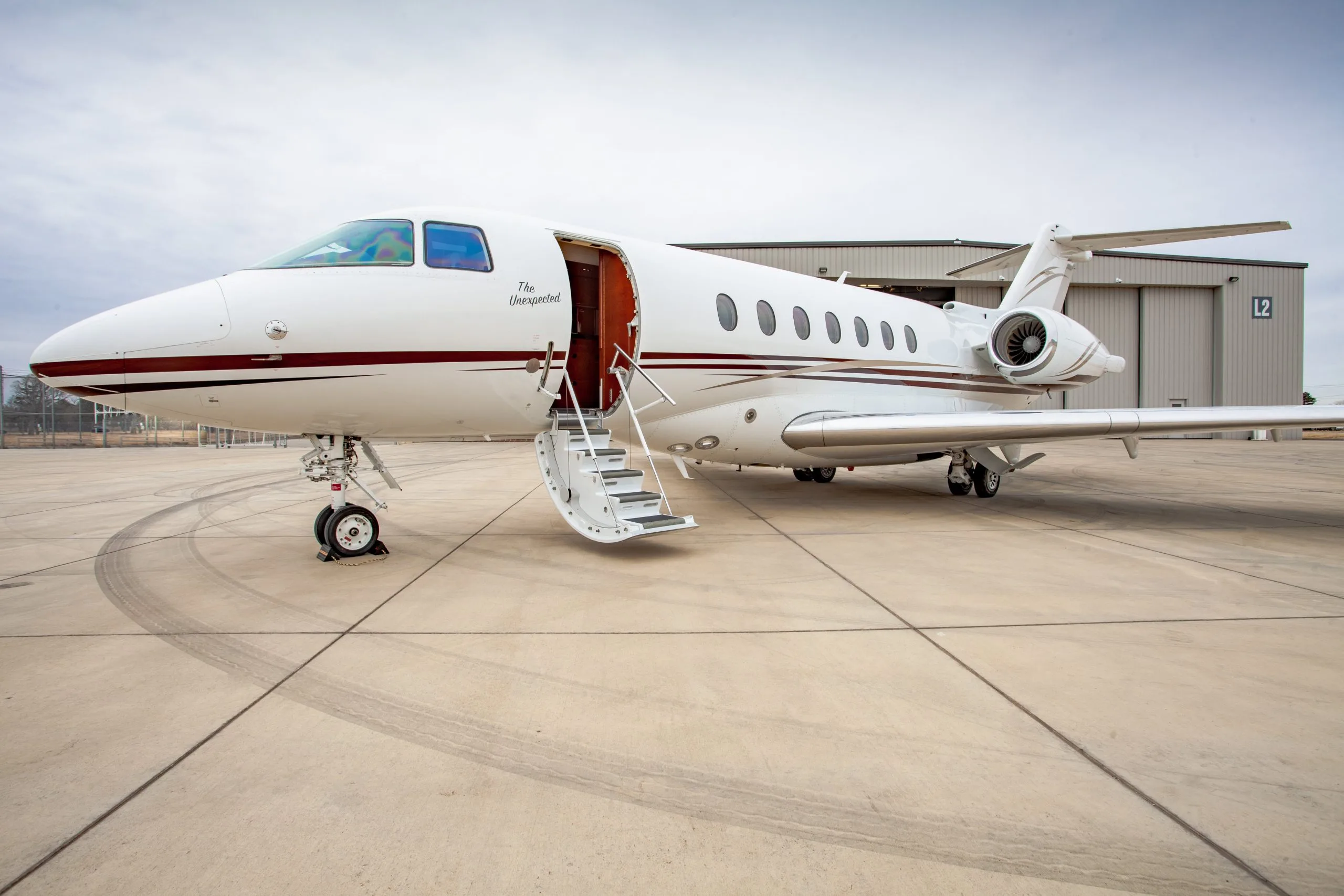 A white business jet with a red stripe and 'The Unexpected' text on its side, parked on an airfield with its door open.