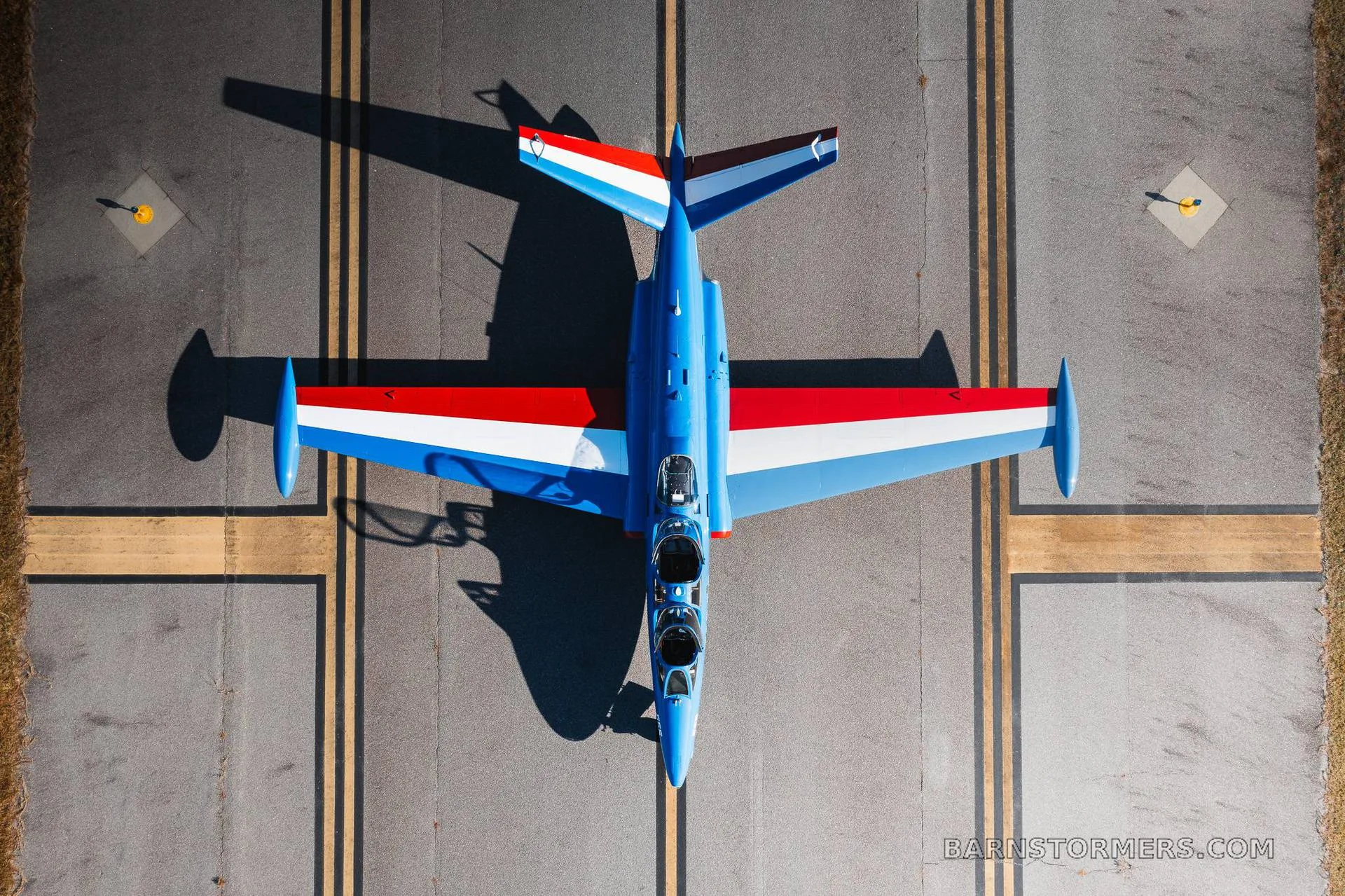 An overhead view of a blue, red, and white Fouga CM.170 Magister jet aircraft parked on an asphalt runway.