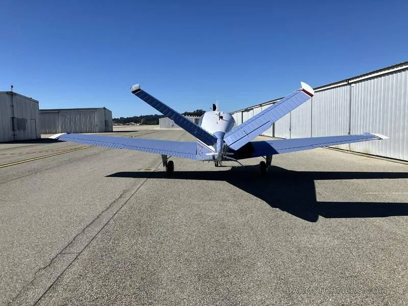 A rear view of a light blue and white single-engine aircraft with its wings folded upwards, parked on an asphalt tarmac.