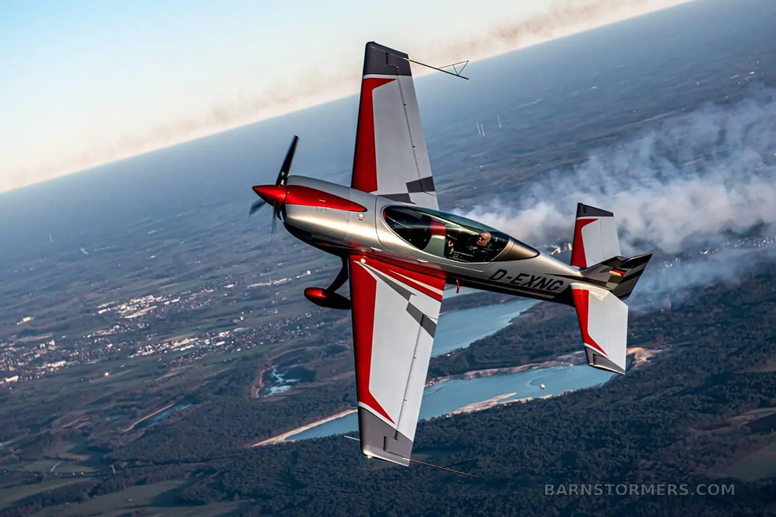 A silver and red Extra 330SC aircraft with registration D-EXNG flies inverted over a landscape.