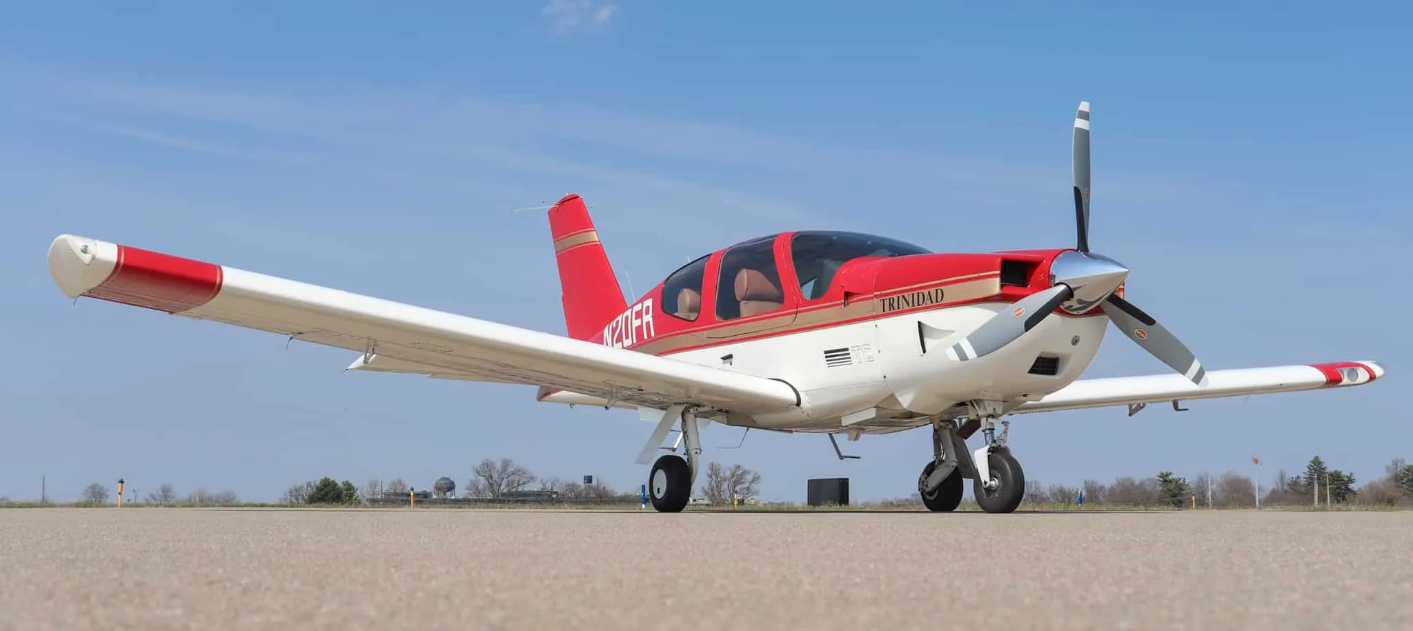 A red and white Socata Trinidad aircraft with gold stripes is parked on an airport tarmac under a blue sky.