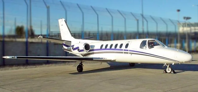 A white business jet with blue and black stripes is parked on an asphalt tarmac with a fence in the background.