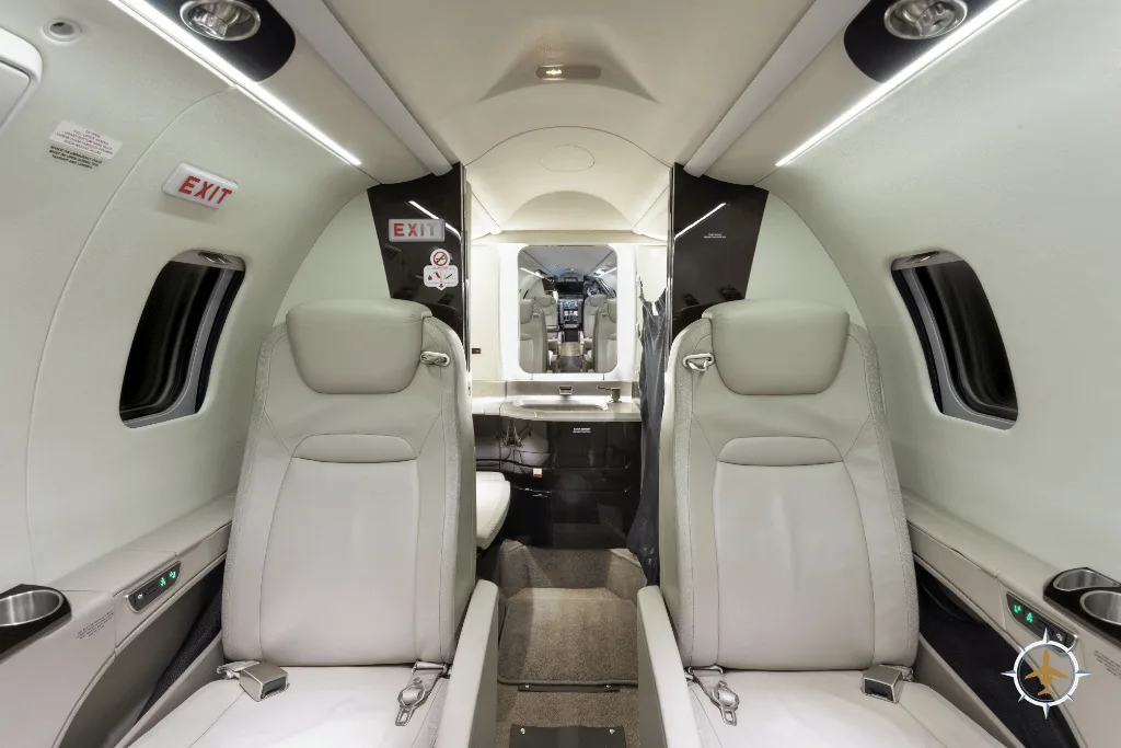 View of a luxurious aircraft cabin with two light-colored leather seats in the foreground and the cockpit visible in the distance.