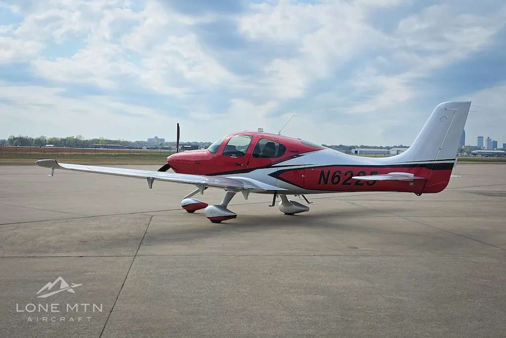 A red and silver Cirrus SR22 aircraft, N6225, parked on an airport apron under a cloudy sky.