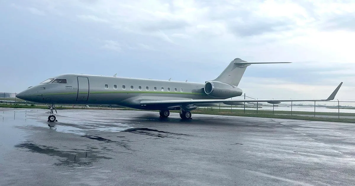 A grey Bombardier Global 5000 business jet with a green stripe on a wet tarmac under a cloudy sky.