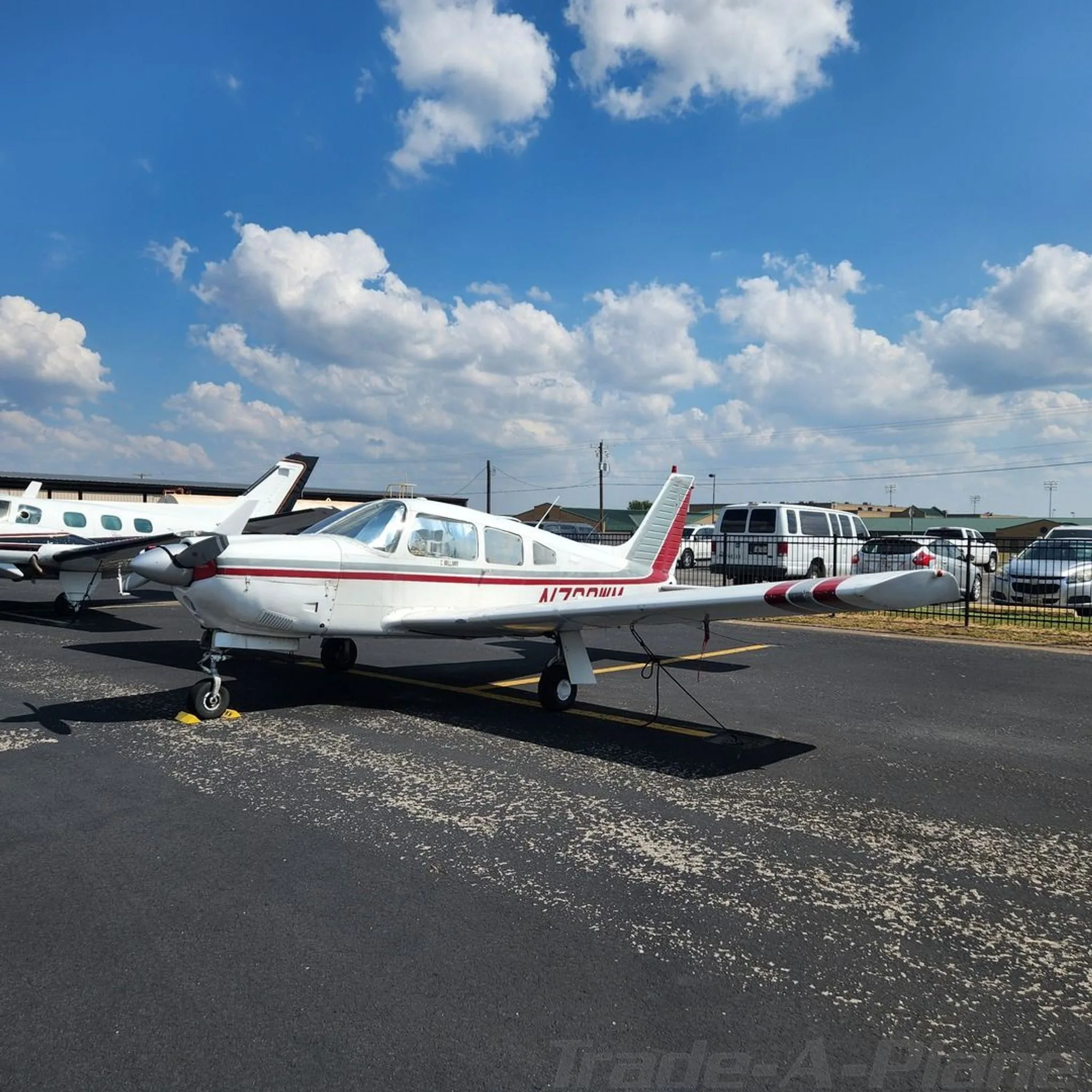 A white Piper Cherokee with red and grey stripes parked on an asphalt ramp under a blue sky with clouds.