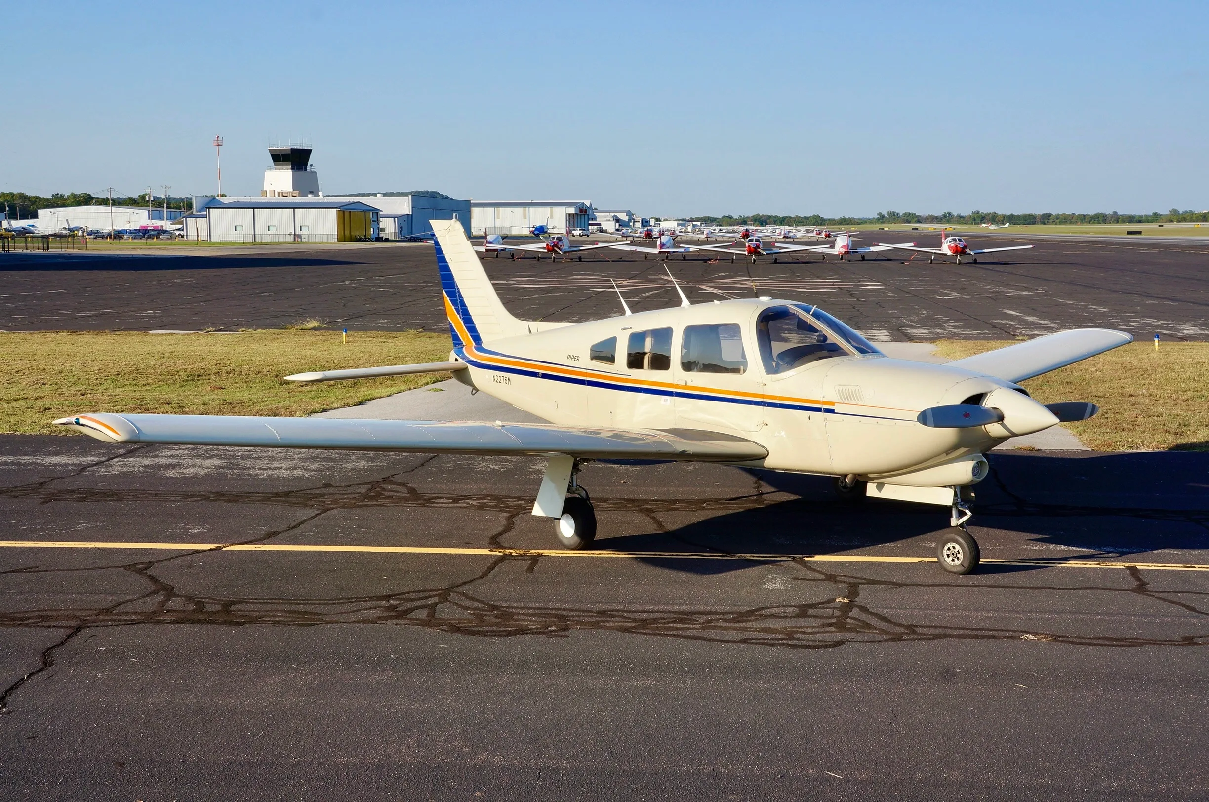 A cream-colored Piper aircraft with blue and orange stripes parked on an asphalt tarmac with a control tower in the background.