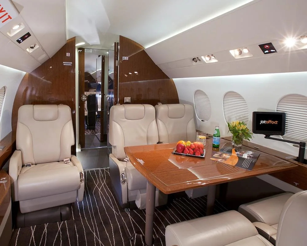 The elegant interior of a business jet cabin, featuring beige seats, a polished wooden table, and a view towards the rear galley area.