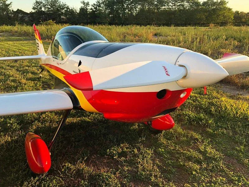 A close-up front view of a white, red, and yellow single-engine aircraft with a propeller on a grassy field.