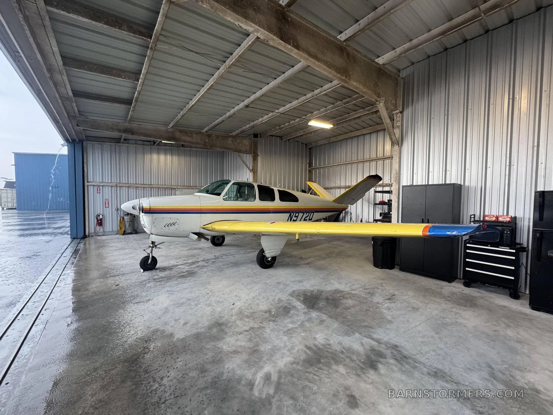 A white and yellow single-engine aircraft with rainbow stripes, viewed from the front-right, inside a hangar with the door open.