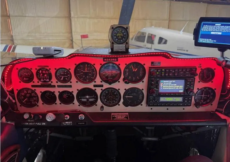The instrument panel of a light aircraft illuminated with red cockpit lighting, showing analog gauges and a radio stack, with another aircraft visible in the background.