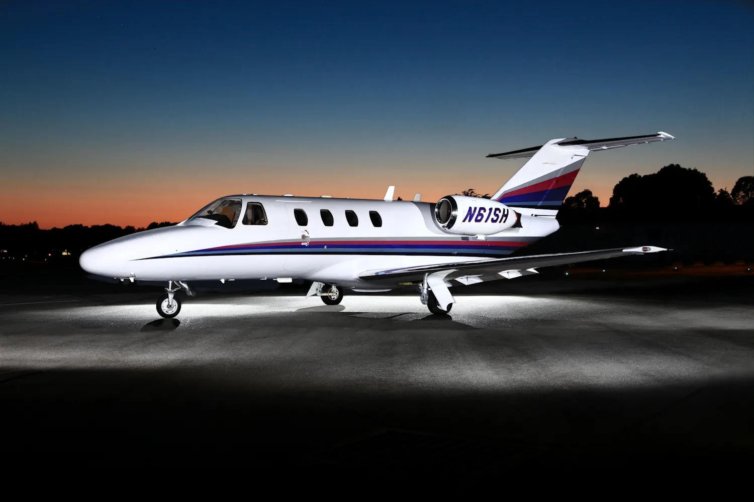 A white private jet with red, blue, and grey stripes on the fuselage and tail, illuminated on an airfield at dusk.