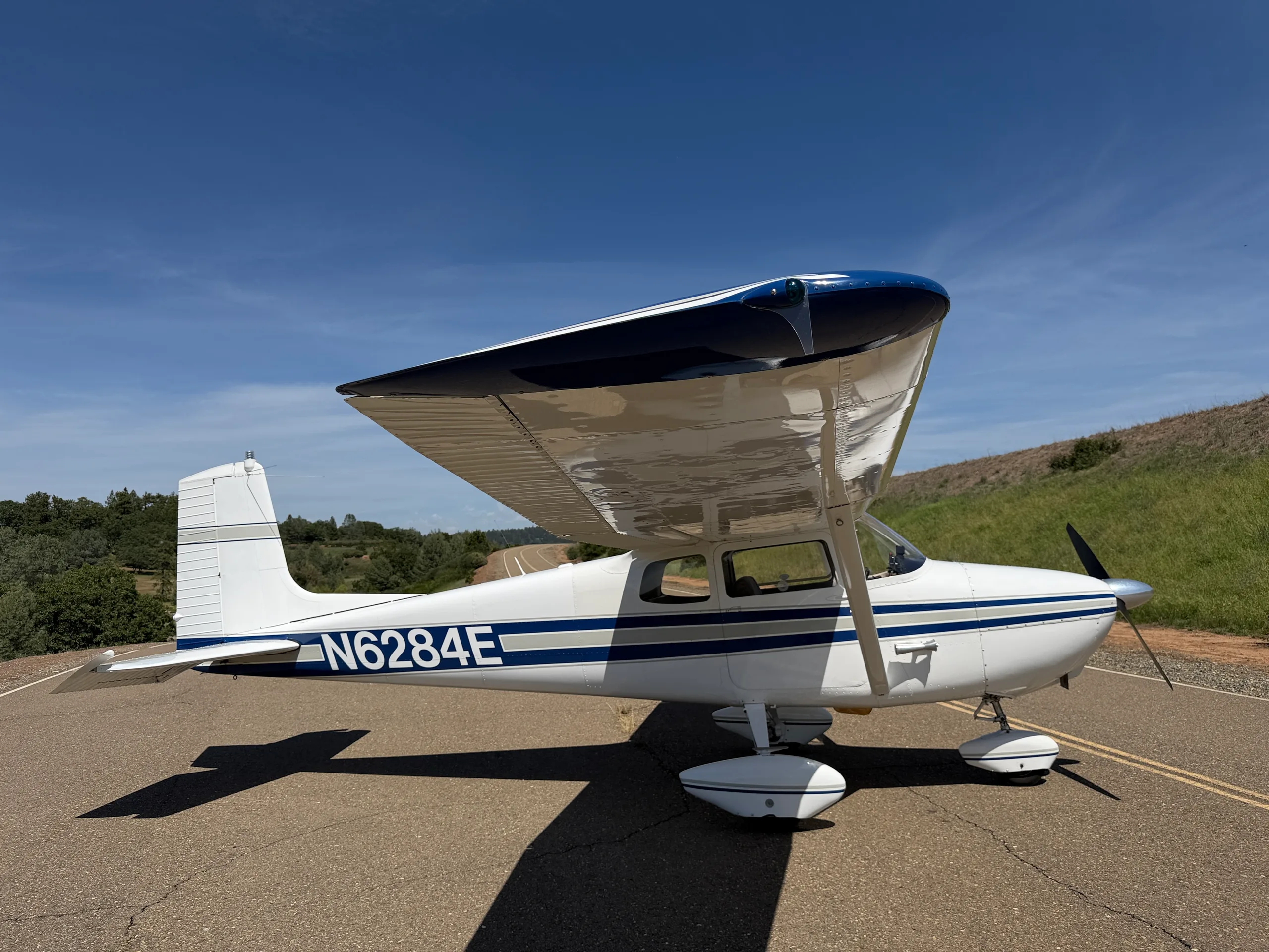 A white single-engine high-wing aircraft with blue and grey stripes, registration N6284E, parked on an asphalt surface under a clear blue sky with green hills in the background.