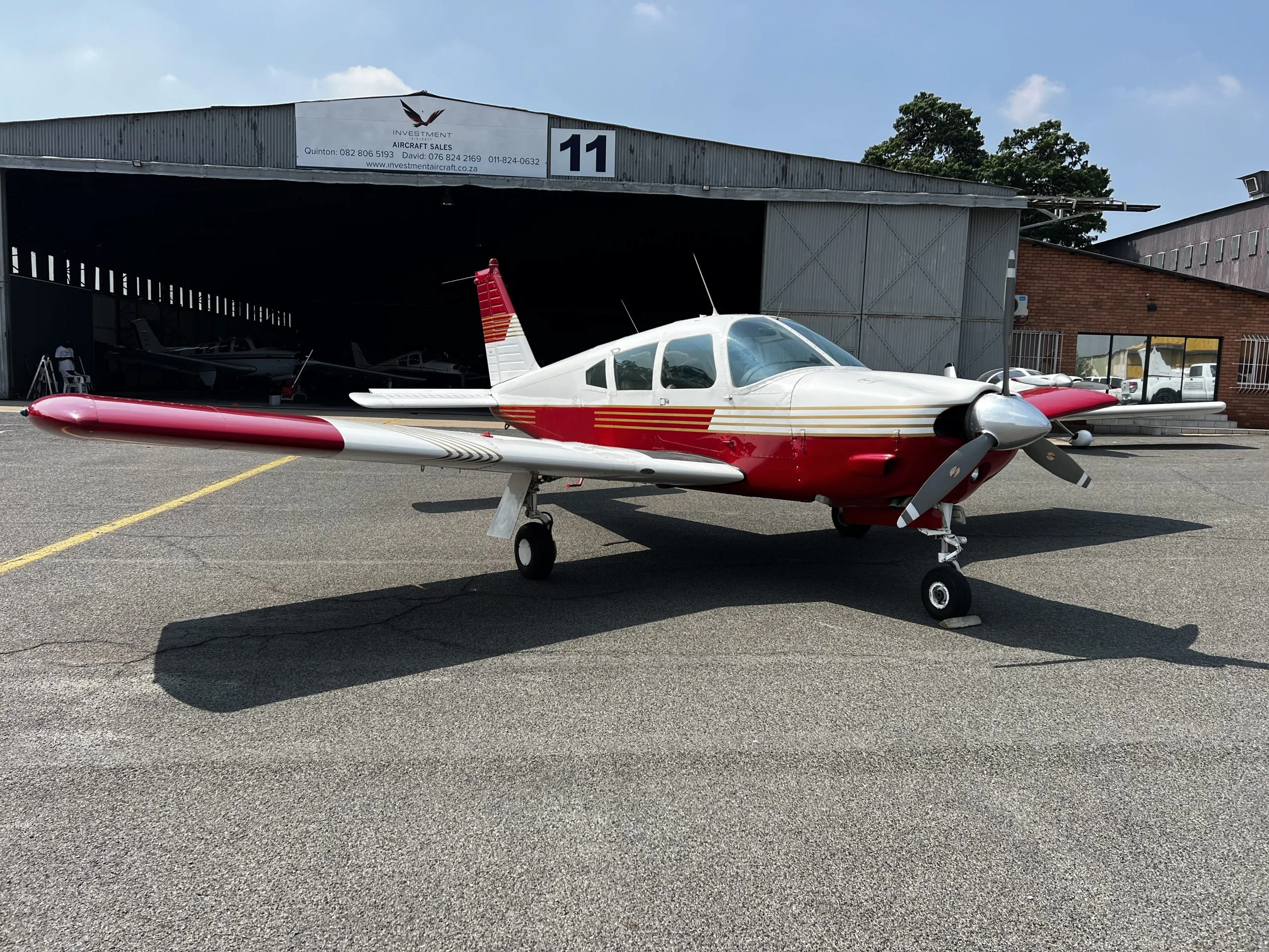 A white and red single-engine propeller aircraft with gold stripes parked on an airfield in front of a hangar.