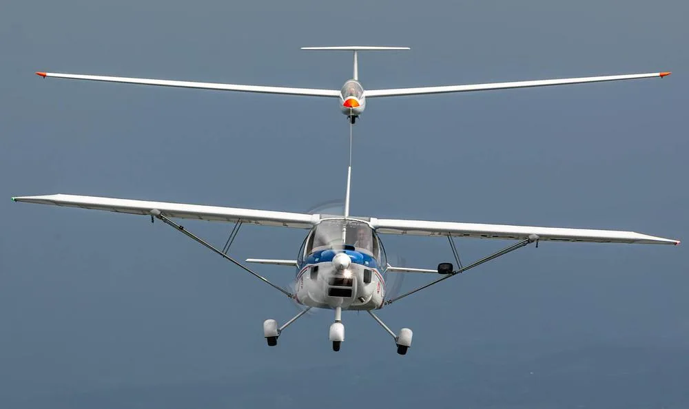 A white and blue tow plane pulls a white glider against a cloudy sky, viewed from the front.
