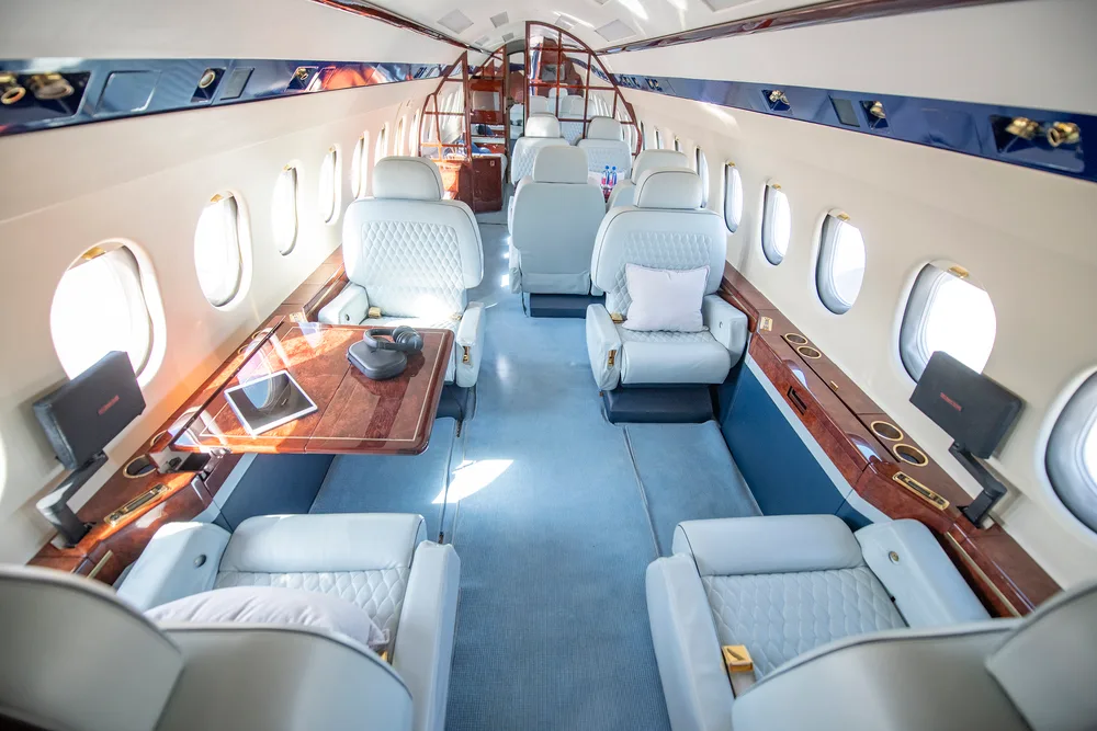 The luxurious cabin interior of a private jet, featuring light grey quilted leather seats, a polished wooden table, and blue accents.