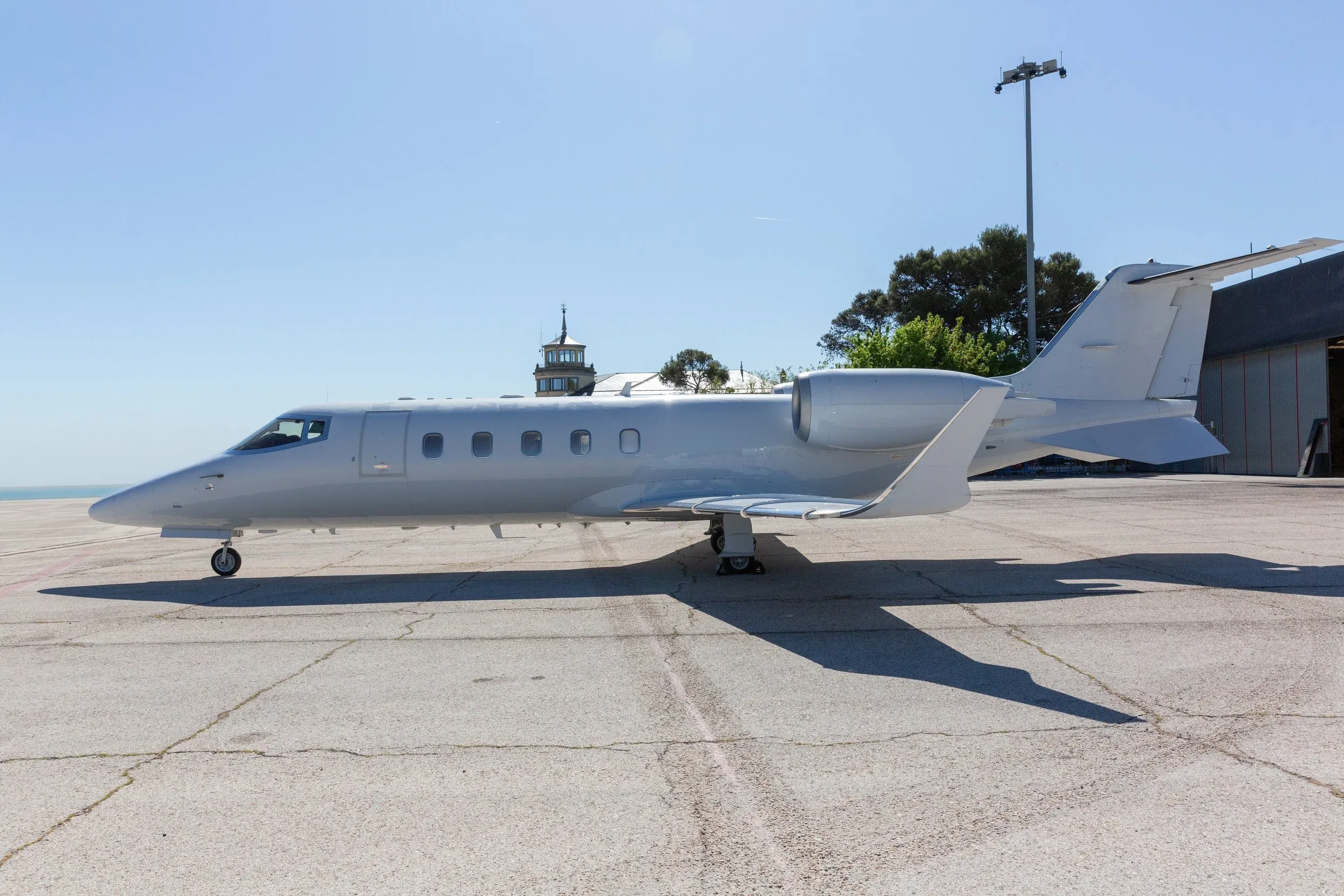 A white business jet, possibly a Learjet, parked on an airfield with a control tower and trees in the background.