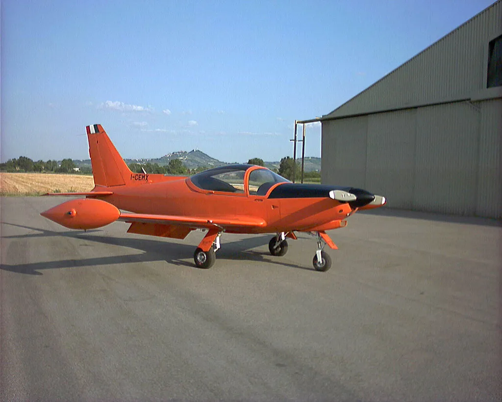 An orange and black single-engine aircraft, possibly a Falco F.8L, is parked on an asphalt apron next to a hangar under a clear sky.