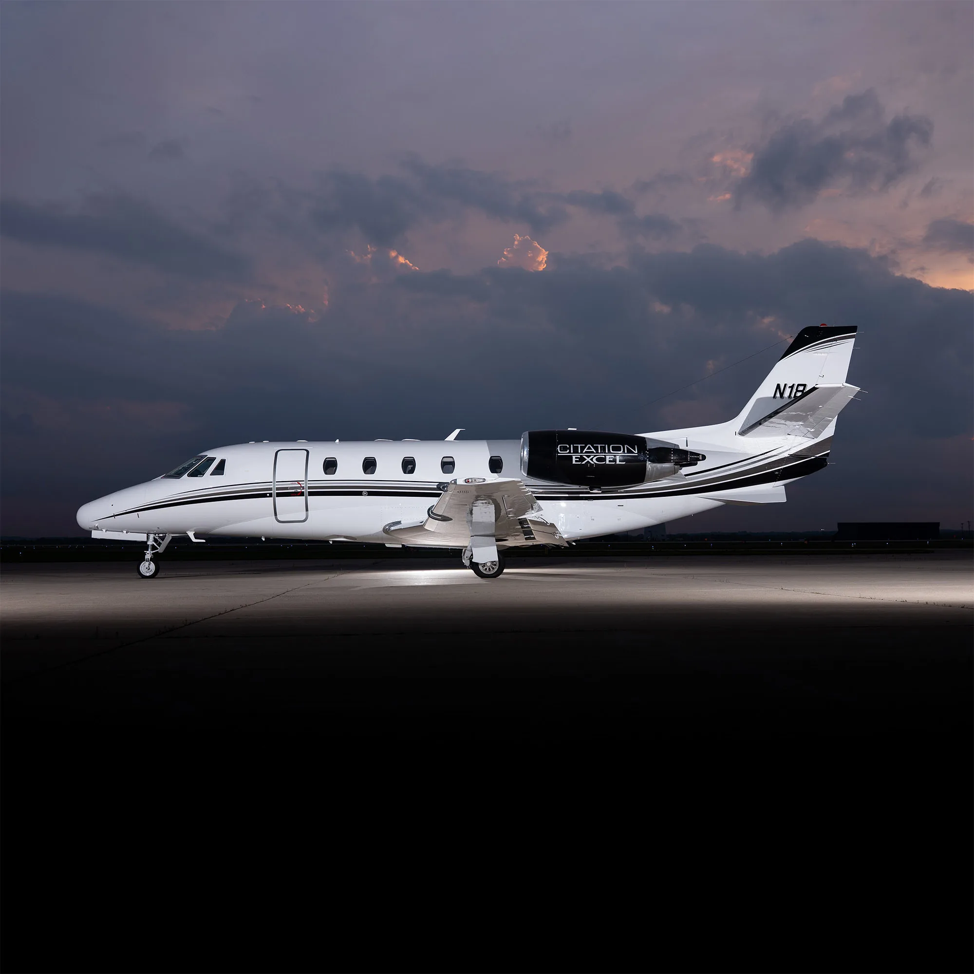 A white Citation Excel business jet with black and white stripes on the tail and engines, parked on a tarmac at dusk.